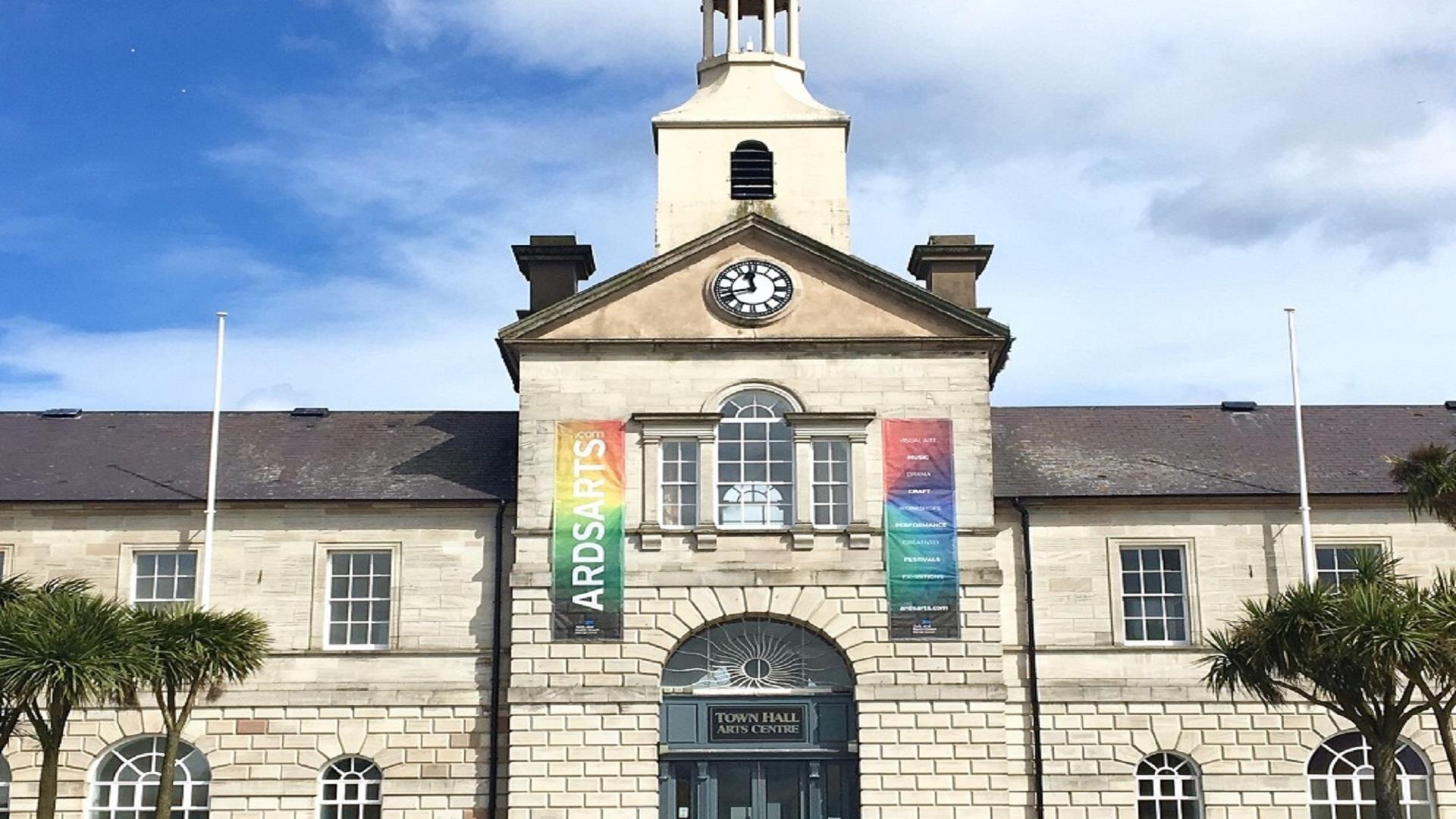Photograph of the front exterior building of Ards Arts Centre previously the Town Hall, with backdrop of blue sky and two banners flanking the front w
