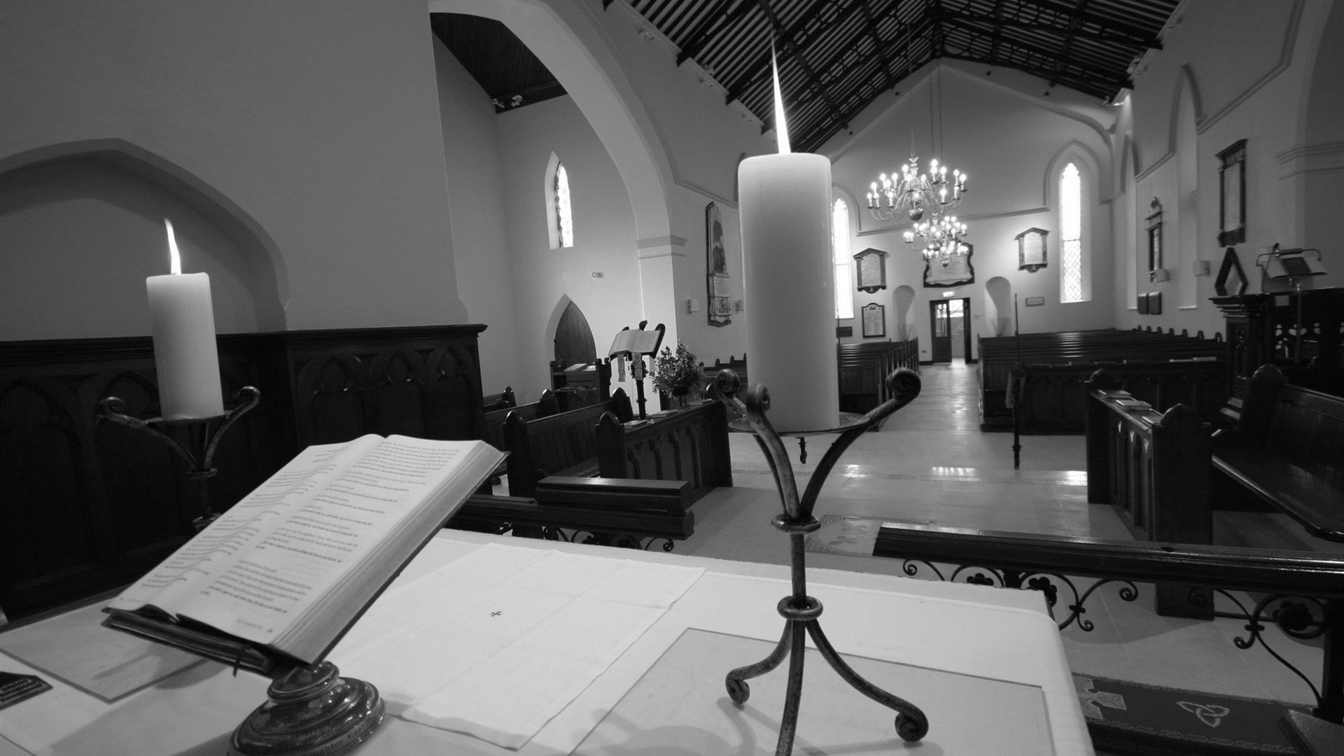 Black and white photo of from the alter of the church showing the Bible and lit candlesticks overlooking the pews
