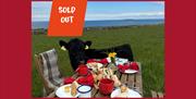 A calf standing beside a picnic table laid with food in a field overlooking the sea with the text 'Sold out'