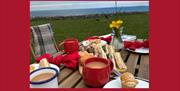 A picnic table set out with food, plates, cutlery and tea, in the middle of a field overlooking the sea