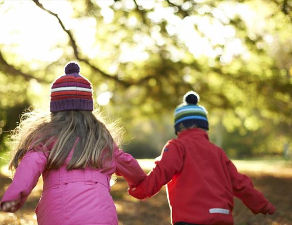 Photo of two children with woolly hats on running through the autumn leaves of the park