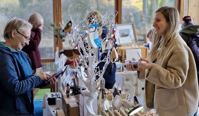 A craft stall at a North Down Craft Collective Fair at WWT Castle Espie.  A visitor is chatting to a local artist.