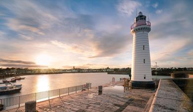 image of lighthouse and pier with sunset over the water in the background.