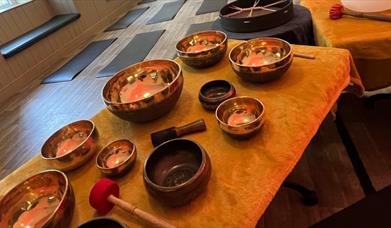A collection of singing bowls laid out on a table. Yoga mats are set out on the floor in the background.
