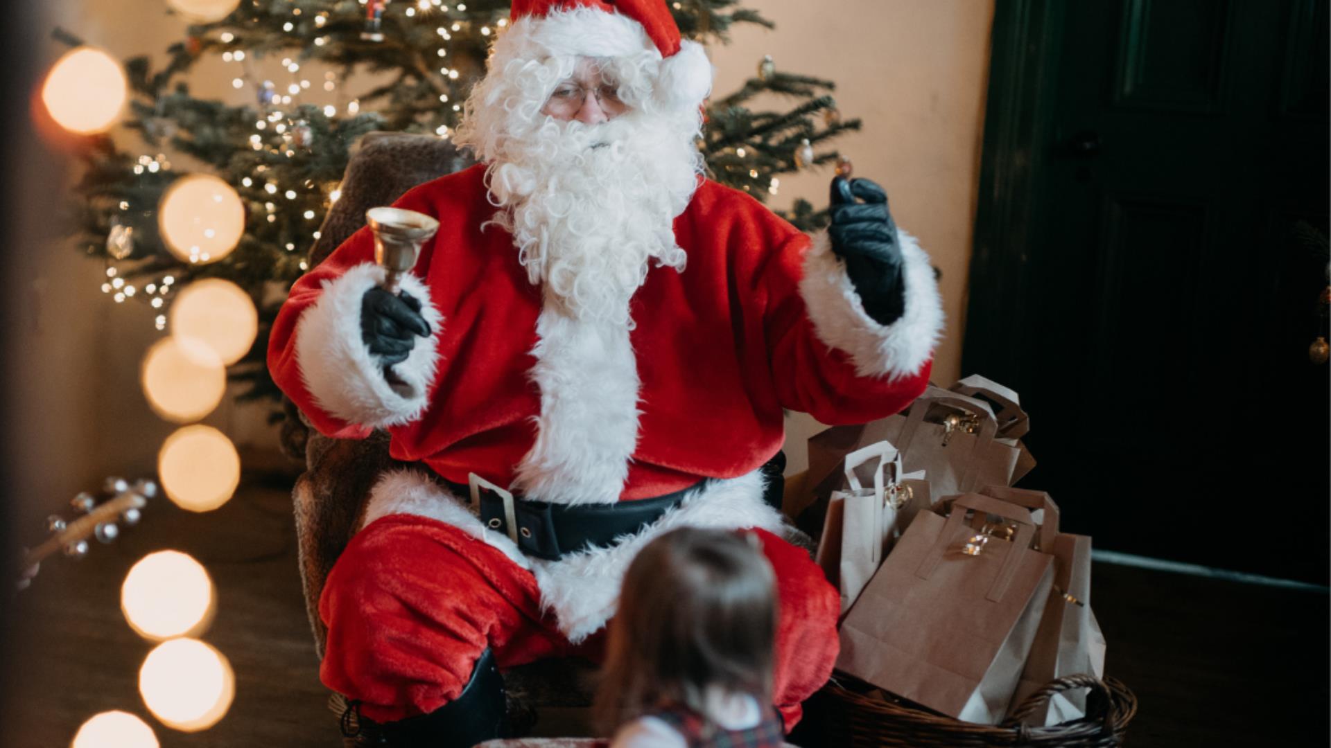 Santa Claus seated beside a Christmas tree, greeting a child in a festive indoor setting with wrapped gifts nearby.
