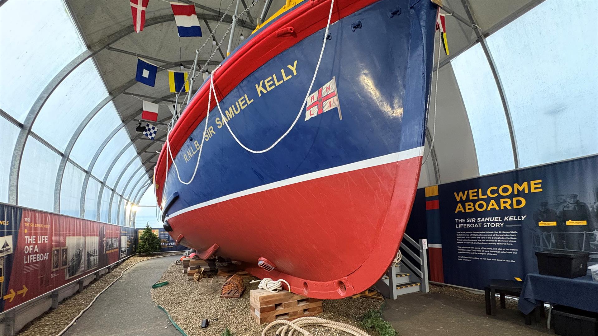 Large red and blue lifeboat in the maritime exhibition space, with nautical flags overhead and interpretive displays alongside.