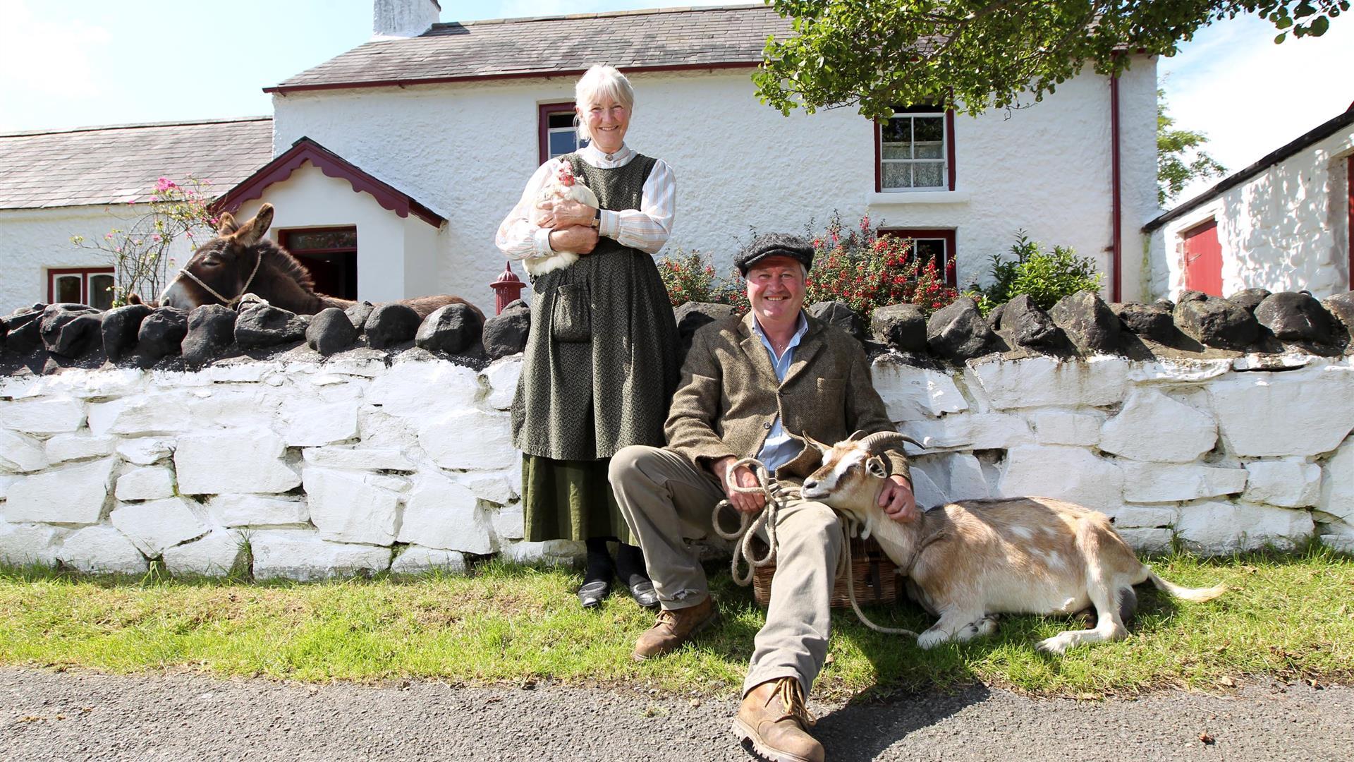 a photograph of two people dressed in old clothing with a goat and a chicken outside some cottages
