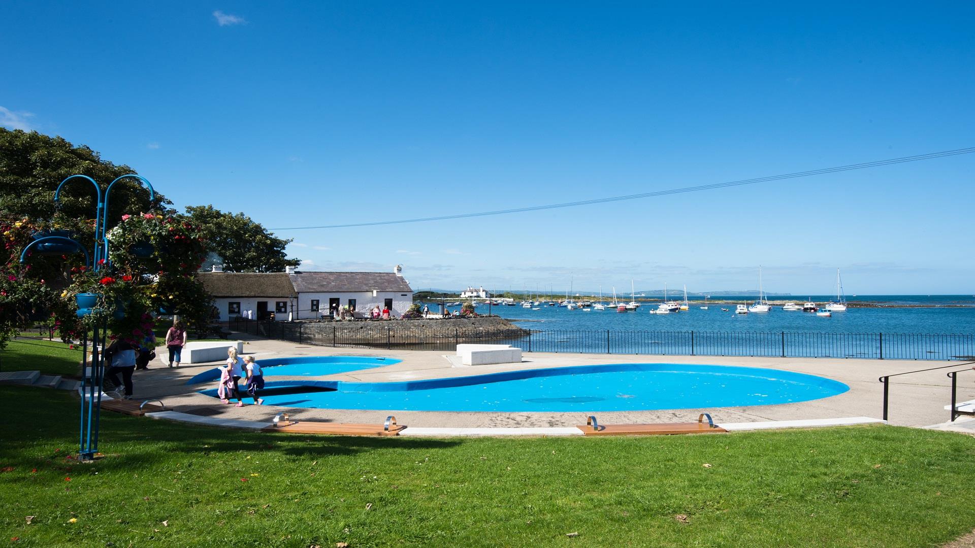 A photo of Cockle Row Cottages taken from a distance to include the seasonal paddling pool in shot on a bright sunny day
