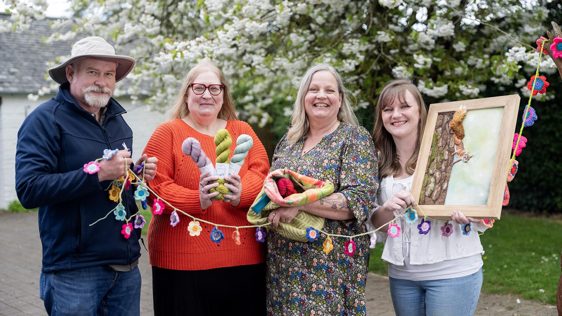 Group of festival organising team and vendor holding crocheted flower bunting and product samples in front of a flowering tree