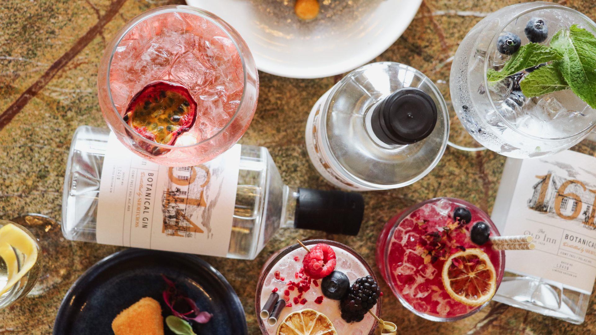 Gin bottles with 1614 gin labels with glasses filled with multiple drinks and cocktails, food plates including brie and fish dish