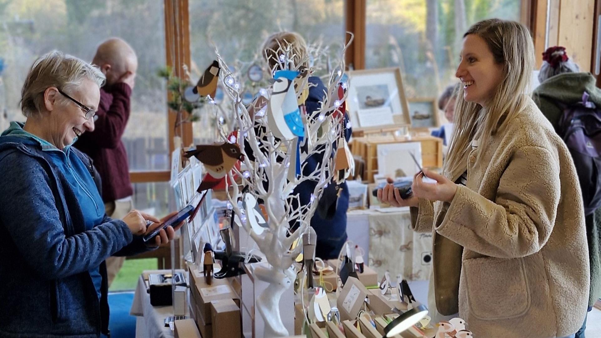 A craft stall at a North Down Craft Collective Fair at WWT Castle Espie.  A visitor is chatting to a local artist.