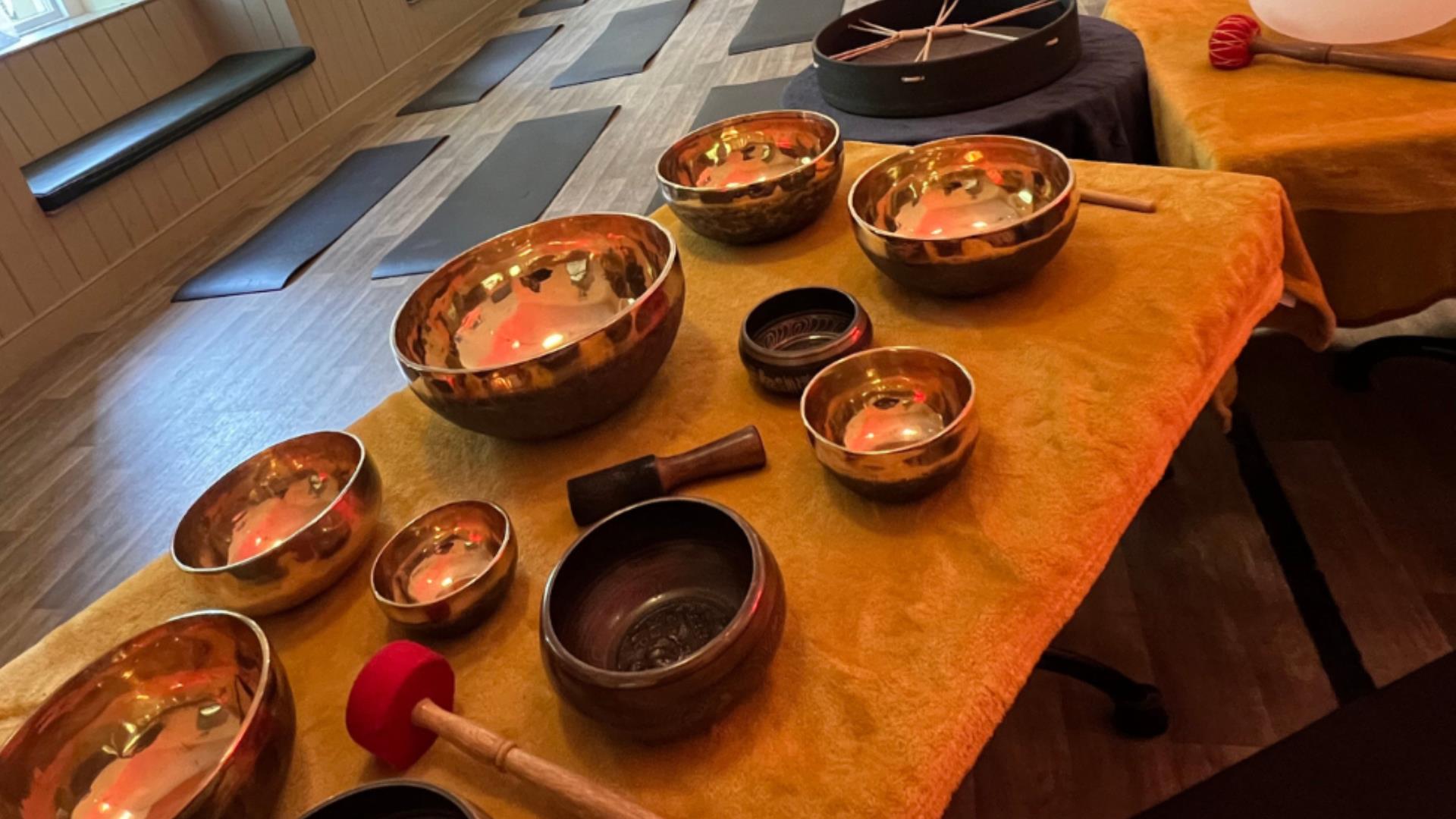 A collection of singing bowls laid out on a table. Yoga mats are set out on the floor in the background.
