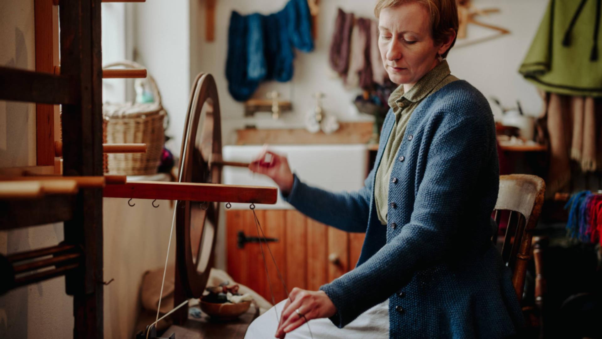 A lady in a blue top and white skirt sitting down and weaving wool