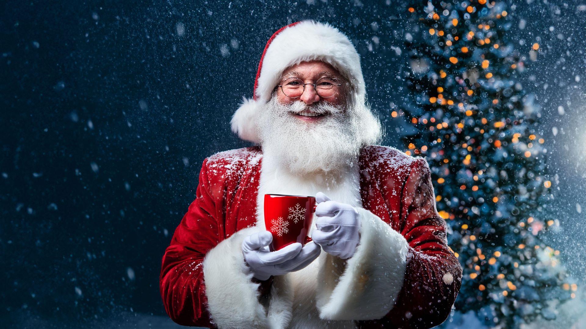 Santa standing in front of a Christmas tree and holding a festive mug. The background is snowy.