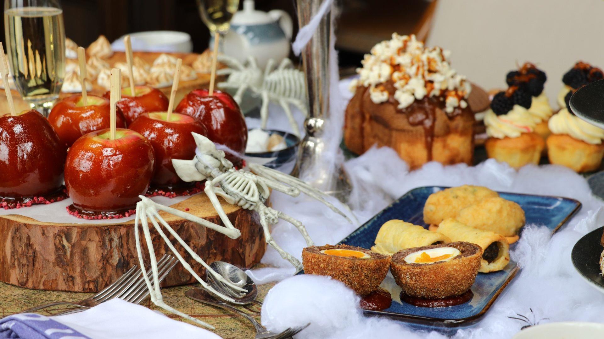 Table set up with food including toffee apples, scotch eggs, sausage rolls, cake and cupcakes with halloween decor