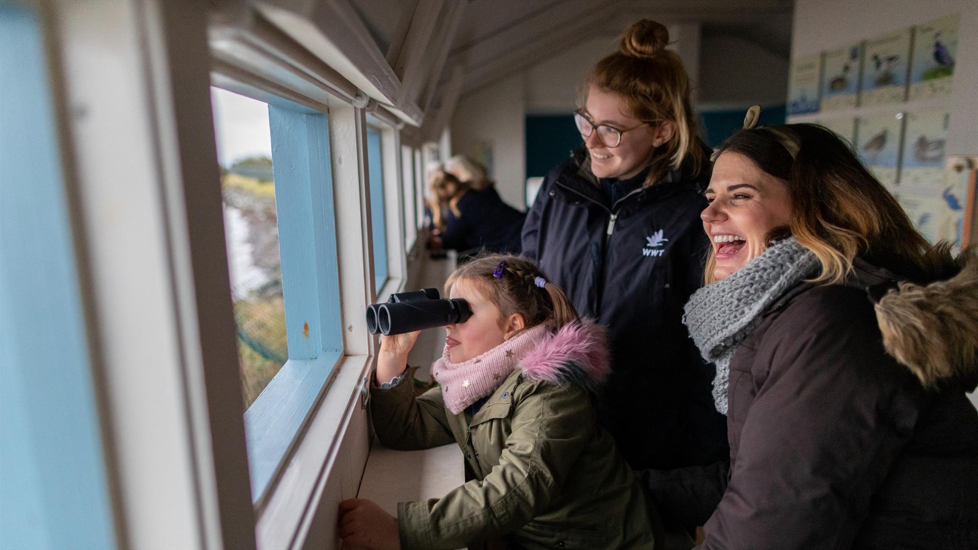 Child looking through binoculars, sitting with her mother and a member of WWT staff in a bird hide.