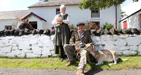 a photograph of two people dressed in old clothing with a goat and a chicken outside some cottages
