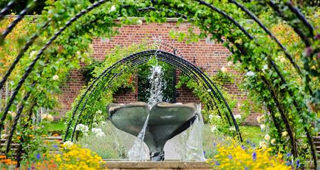 a photograph of a water fountain in the bangor walled garden surrounded by metal arches and colourful flowers