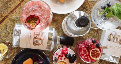 Gin bottles with 1614 gin labels with glasses filled with multiple drinks and cocktails, food plates including brie and fish dish