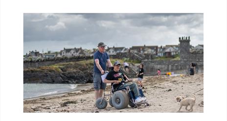 Image showing a young adult on Groomsport Beach seated in an all-terrain beach wheelchair. He is accompanied by a male friend pushing the chair and is