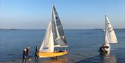 Two small sailing boats being launched from a slipway into calm water, with people preparing boats on the shore.