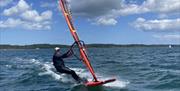 A windsurfer gliding across the water under a blue sky, with a colourful sail and distant shoreline.
