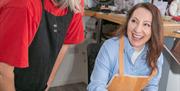 One lady hammering a ring on a ring mandrel, being supervised by a goldsmith with her googles on.  both ladies smiling and looking like they are havin