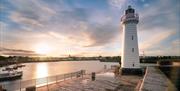 image of lighthouse and pier with sunset over the water in the background.
