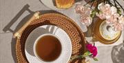 A cup of tea on a woven tray, accompanied by brown sugar cubes, slices of cake on a decorative plate, and a bouquet of pink carnations—all beautifull