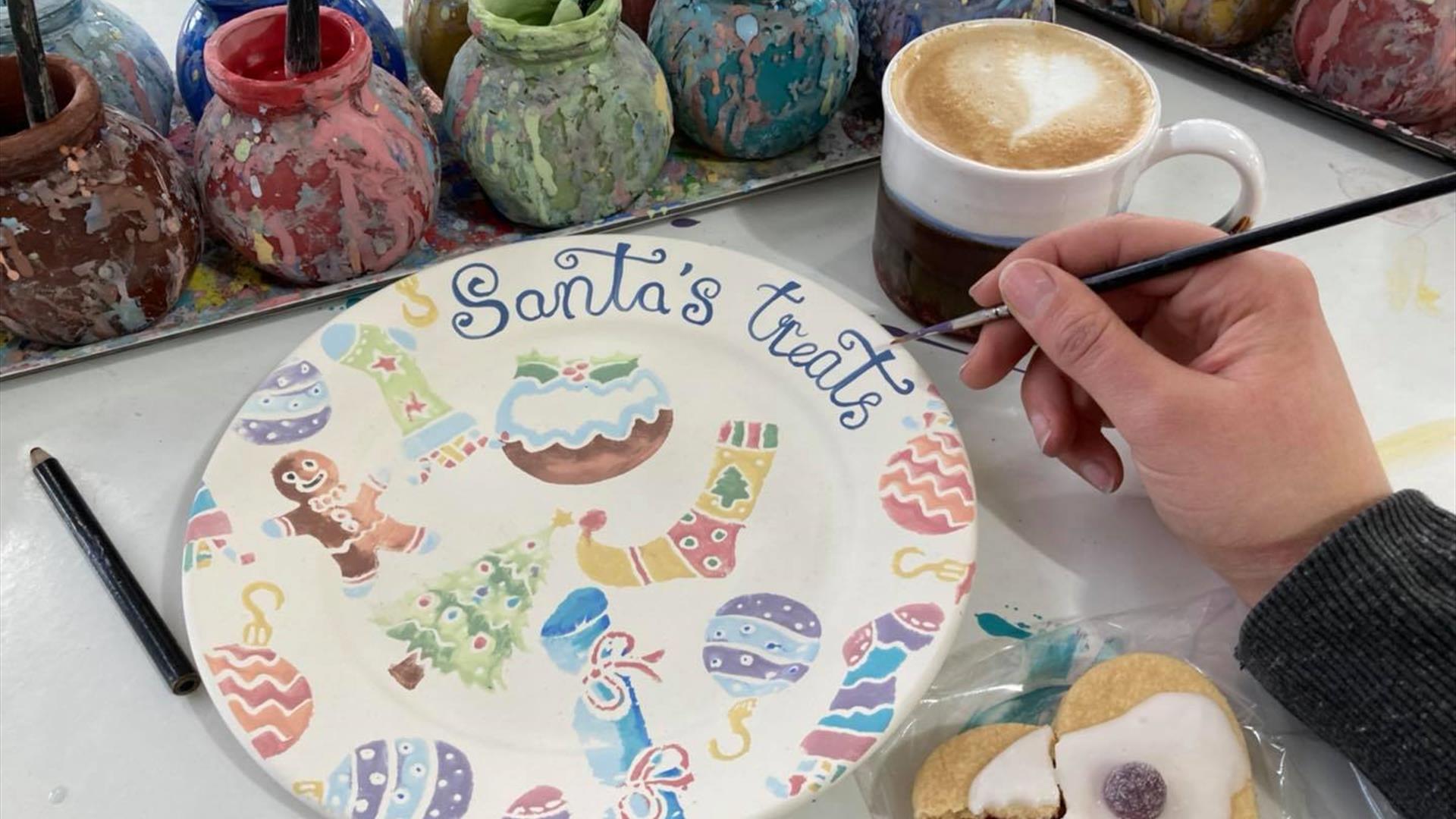 A hand sponged side plate with Santa's Treats written on it, an arm holding a paint brush, a mug of coffee, and a half eaten german biscuit