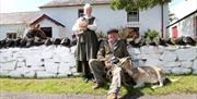 a photograph of two people dressed in old clothing with a goat and a chicken outside some cottages
