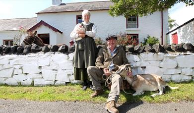 a photograph of two people dressed in old clothing with a goat and a chicken outside some cottages