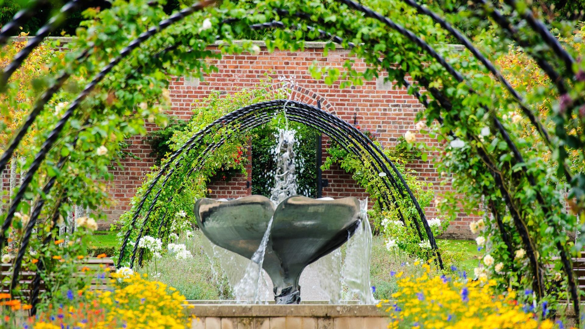 a photograph of a water fountain in the bangor walled garden surrounded by metal arches and colourful flowers