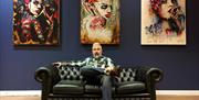 Photo of artist Terry Bradley seated on couch in front of three of his paintings hanging on display