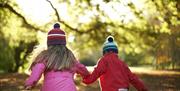 Photo of two children with woolly hats on running through the autumn leaves of the park