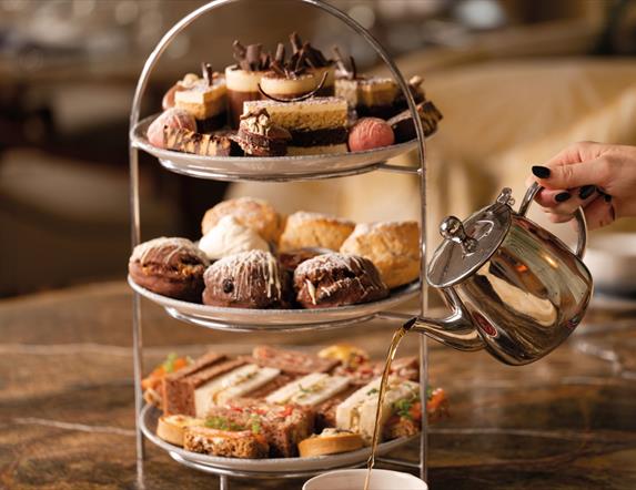 Three tiered selection of savoury and sweet treats, including, sandwiches, scones and desserts. With a hand pouring tea from a teapot into a cup.