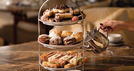 Three tiered selection of savoury and sweet treats, including, sandwiches, scones and desserts. With a hand pouring tea from a teapot into a cup.