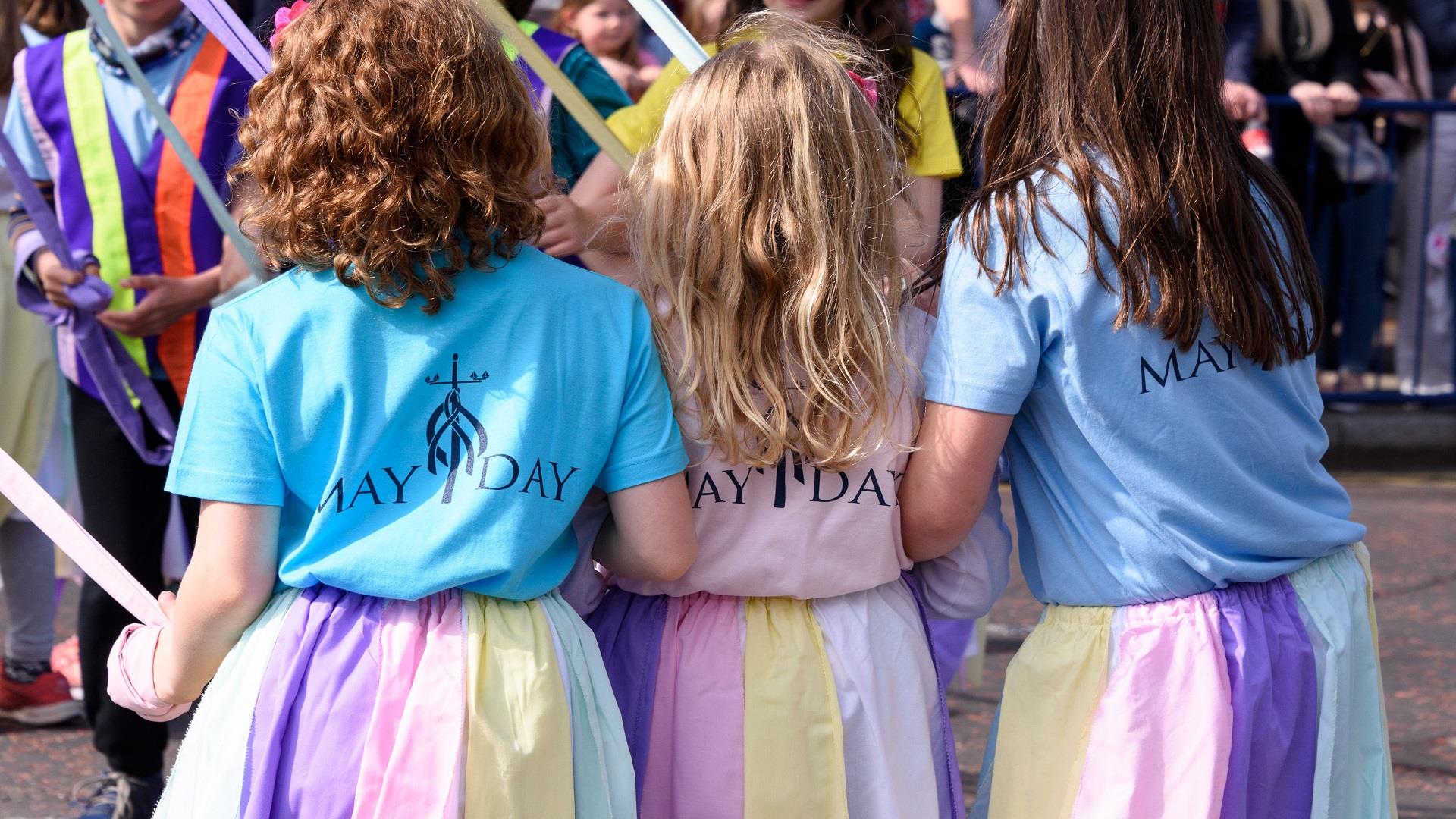 A photo of a trio of little girls taking part in the annual maypole dancing dressed in pastel skirts to match the ribbons of the Maypole