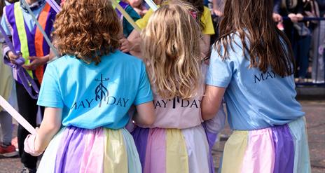 A photo of a trio of little girls taking part in the annual maypole dancing dressed in pastel skirts to match the ribbons of the Maypole