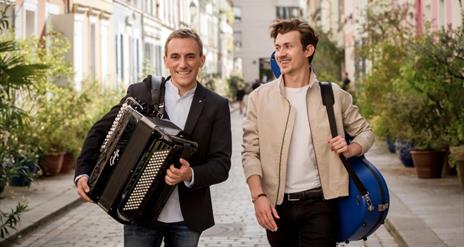 a photo of two men walking through a street holding an accordian and a guitar