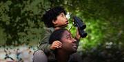 A boy on his father's shoulders, holding binoculars and looking up into the trees.