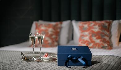 2 flutes of Prosecco and some chocolates on a tray, and a dark blue gift box sitting on a bed.